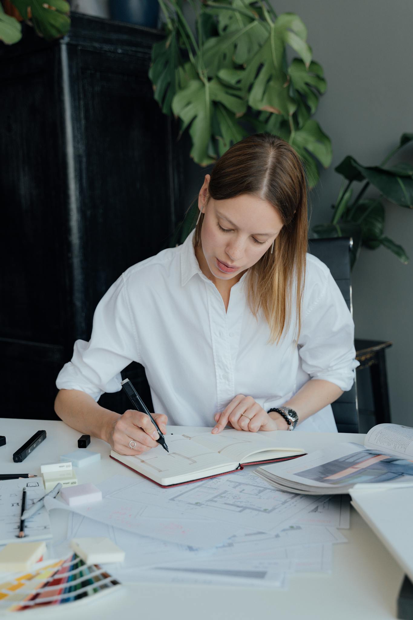 Woman in white shirt working on designs at a desk with various papers and a plant in the background.