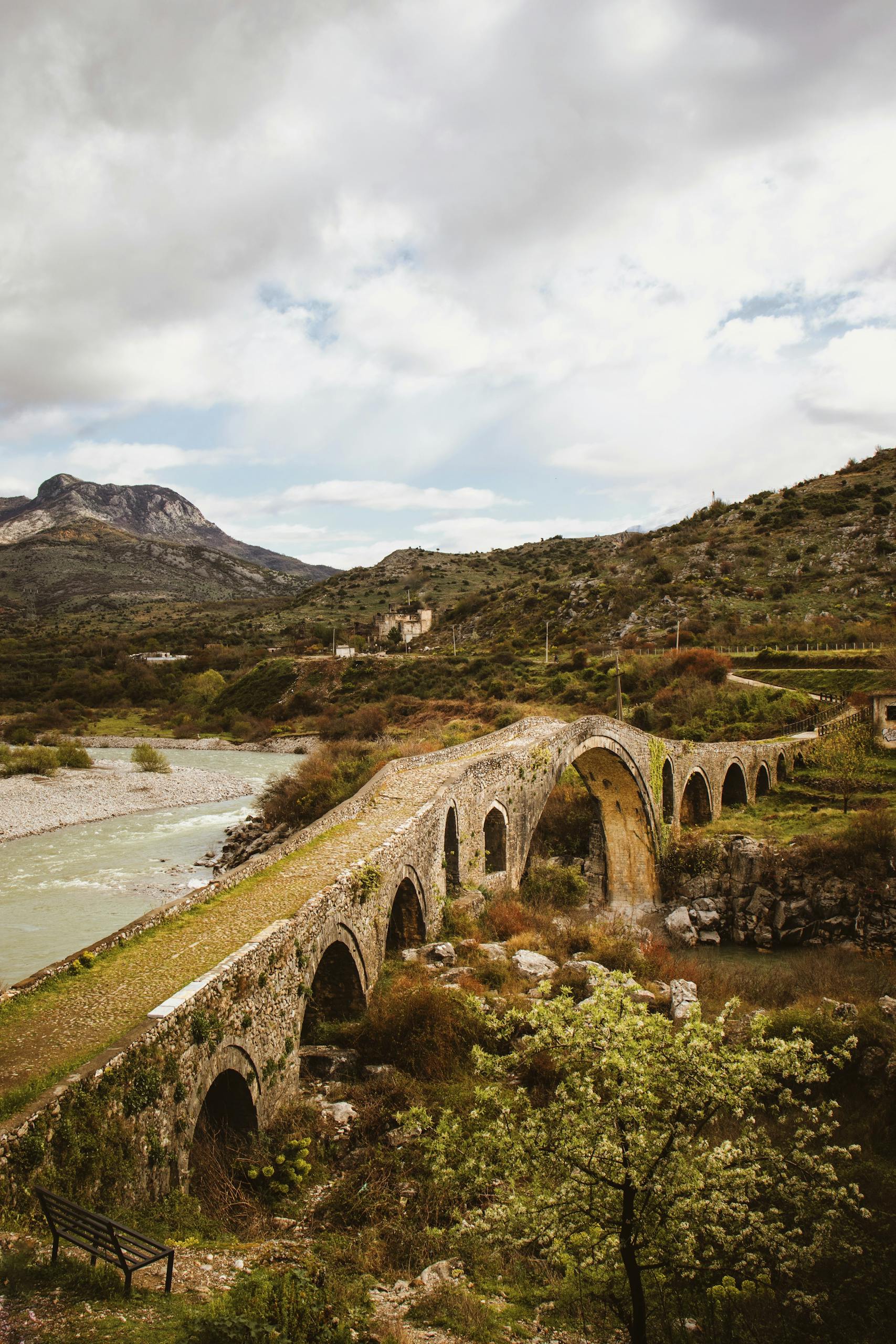 A picturesque view of the historic stone Mesi Bridge over a river in rural Shkodër, Albania.